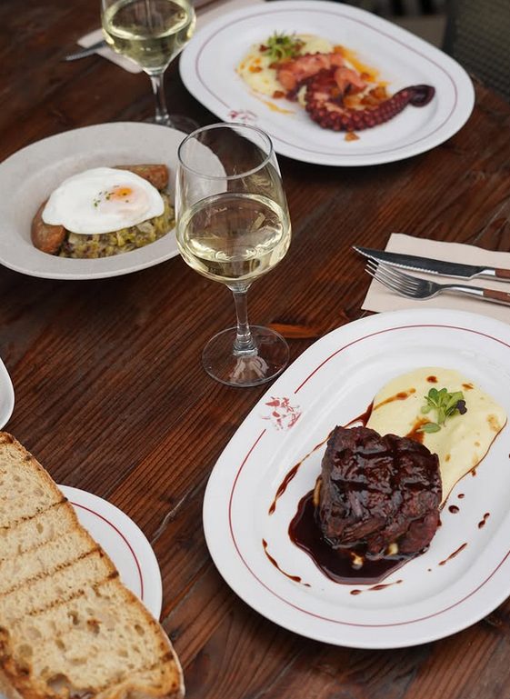 Filet amb puré, pop a la brasa i ou ferrat sobre taula de fusta, amb copes de vi blanc